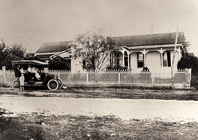 A black-and-white early 1900s photo shows a family sitting in a Model T Ford car in front of a one-story frame house.
