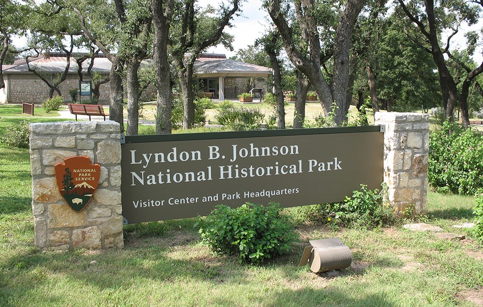 A large brown sign stands in front of a stone building. It reads "Lyndon B. Johnson National Historical Park, Visitor Center and Park Headquarters"