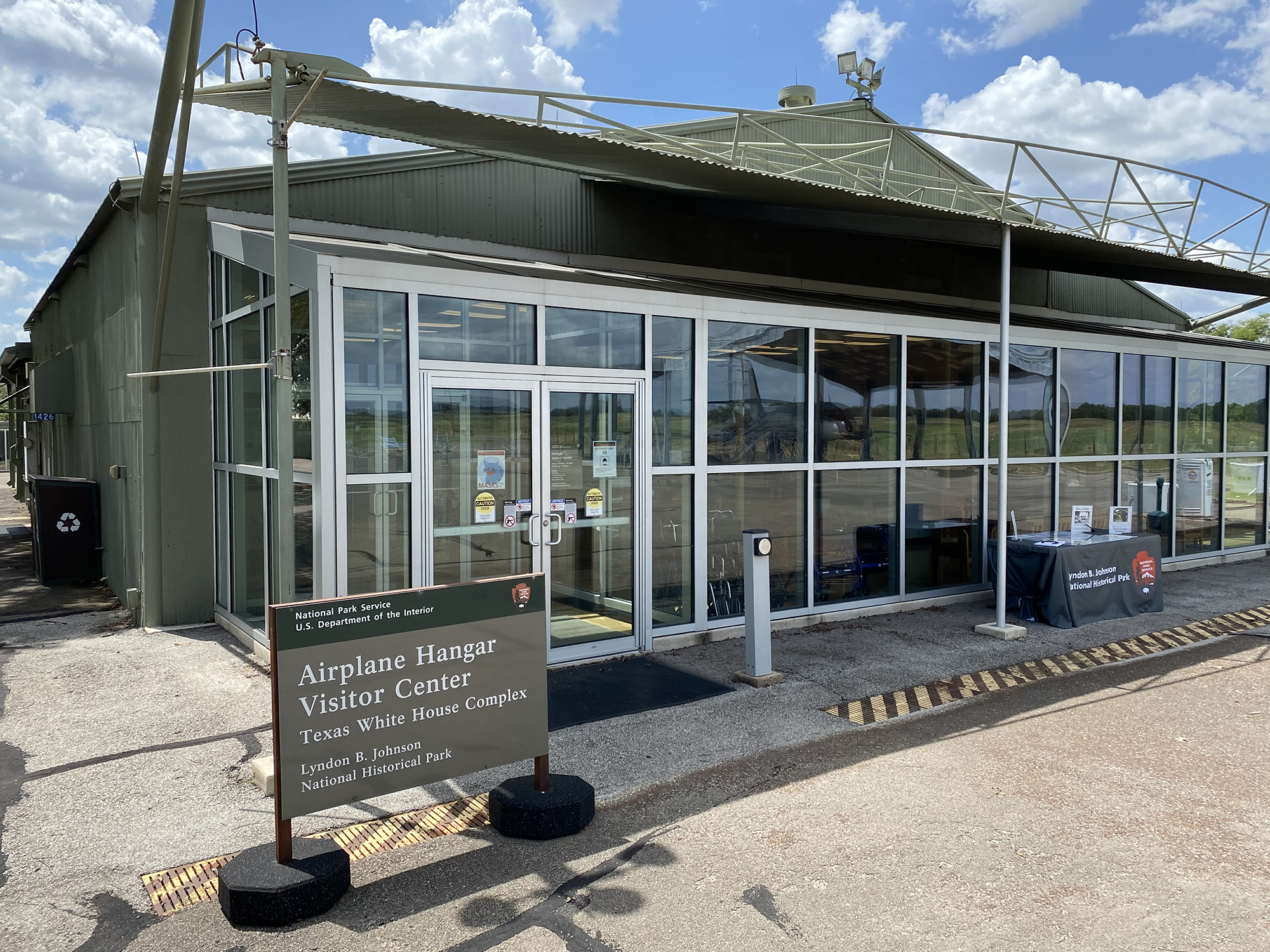 A wall of glass lines the front of the large, green metal airplane hangar. A sign displayed states "Airplane Hangar Visitor Center."