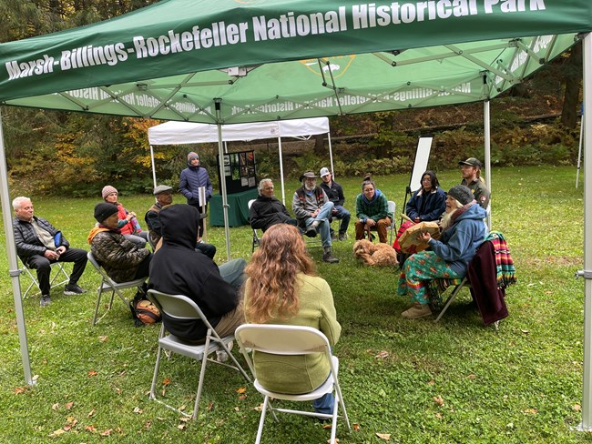 Abenaki person plays hand drum while group of 13 people sits in circle and listen