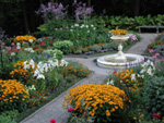 Yellow, white, purple and the occasional red flower distinguish themselves from their greenery in this garden picture. At the center of the garden a round white marble fountain stands.