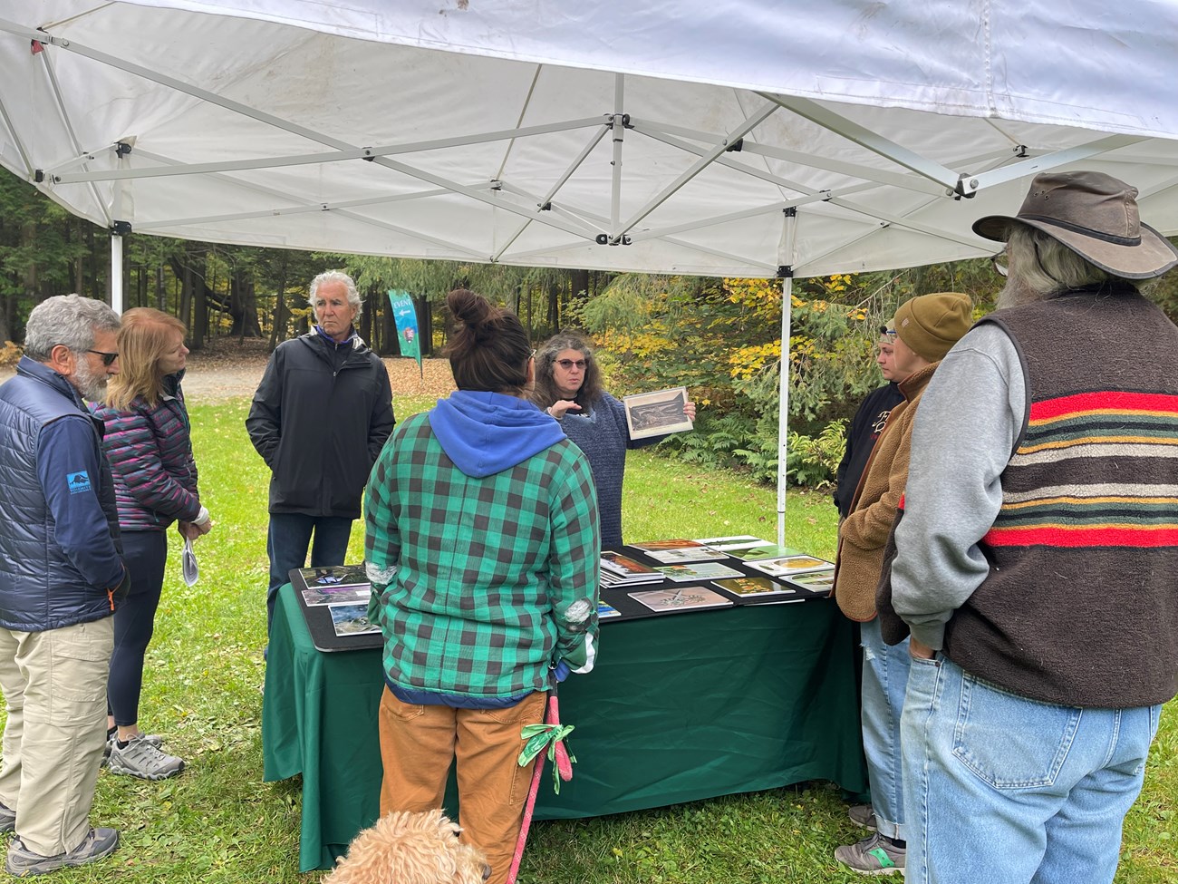 A group of people listen to a woman talk as she shows photos around a table outside.