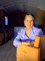 A smiling silver-haired woman in a lavender-colored blouse leans on a wooden altar inside the cool dark of the small chapel.