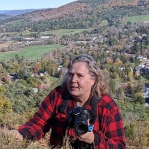 woman stands with camera on top of mountain with view of vermont village behind