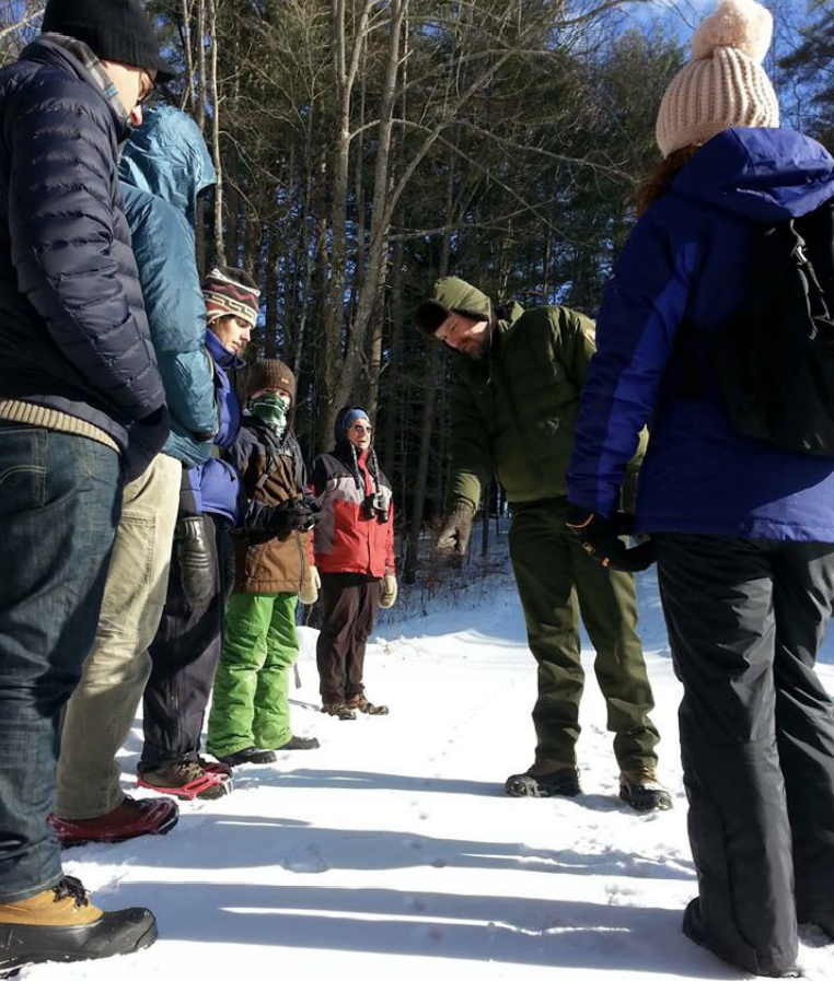 Park Ranger stands outdoors in winter and points at animal tracks surrounded by six people