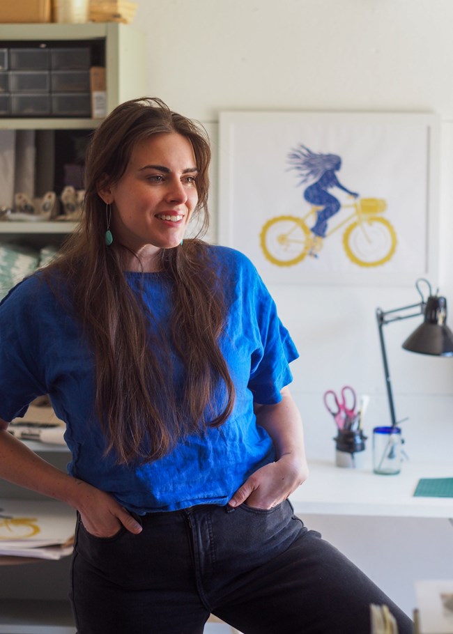 photo of person standing in blue shirt with desk and artwork behind them