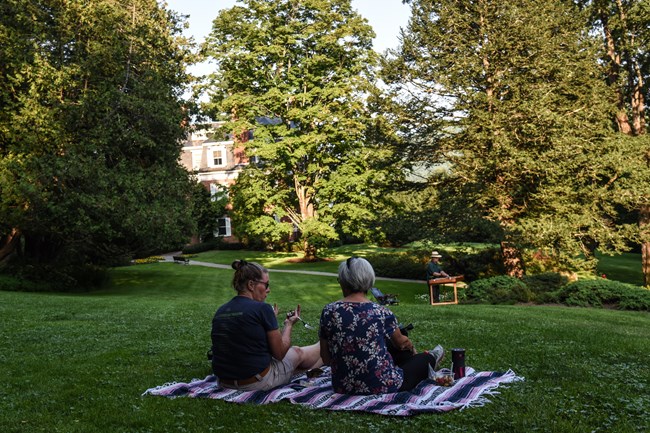 Two visitors sit with their backs to the camera on a picnic blanket overlooking a green lawn