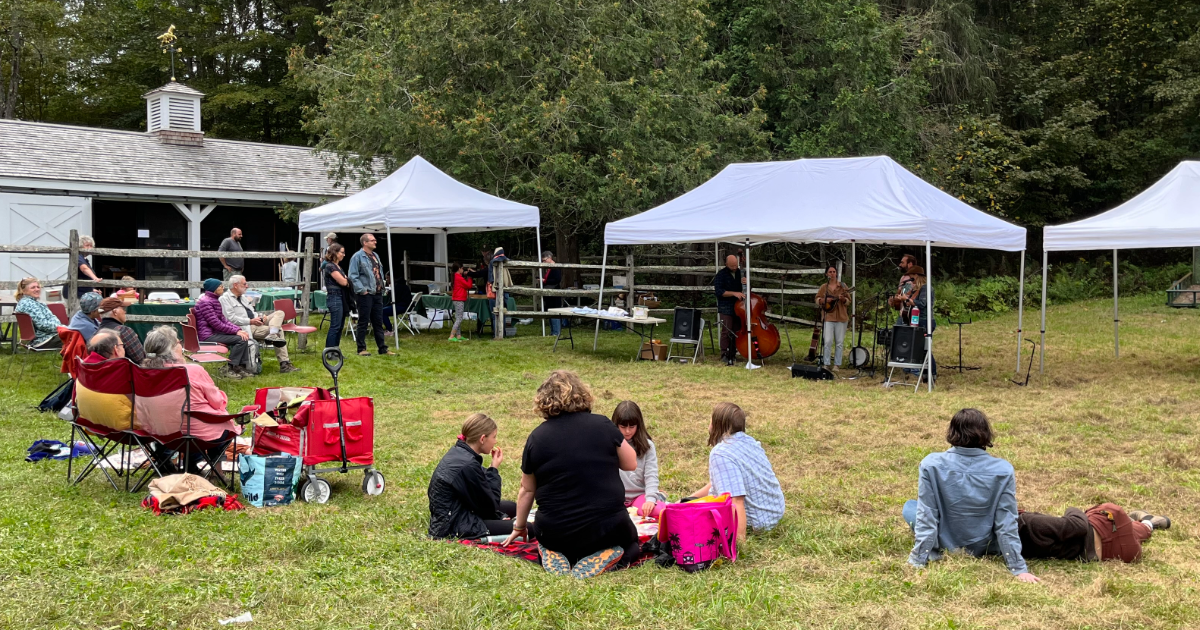 outdoor festival with three white pop up tents, people sit on lawn with blankets and chairs and watch band play under a tent, band includes a standup bass and banjo