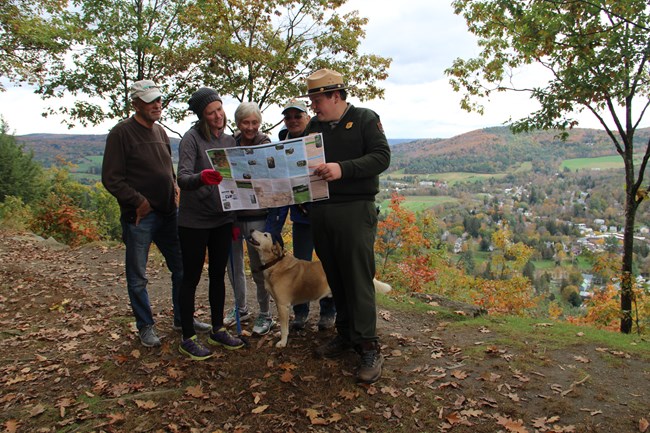 A Park Ranger, volunteer, and three visitors in hiking clothes, and a dog look at a map in front of a fall vista