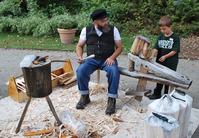A bearded adult man holds a wooden spoon and carving knife while talking to an elementary school aged boy. They are surrounded by wood shavings and wooden equipment.