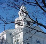 Looking from below, the steeple of this white wooden church is framed by the leafless branches of a nearby tree.