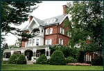 Three story brick mansion with wide porch, white trim, tall chimneys, shrubs and green lawn.