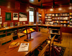Books fill the shelves, and daylight and small lamps warm a large wooden table in the reading area of the bookstore.