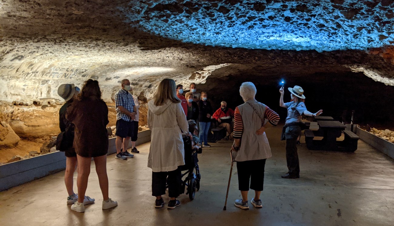 About a dozen people, one with a cane and one in a wheelchair, stand in a semi-circle facing a park ranger. The park ranger points a flashlight directly up at the low cave ceiling.