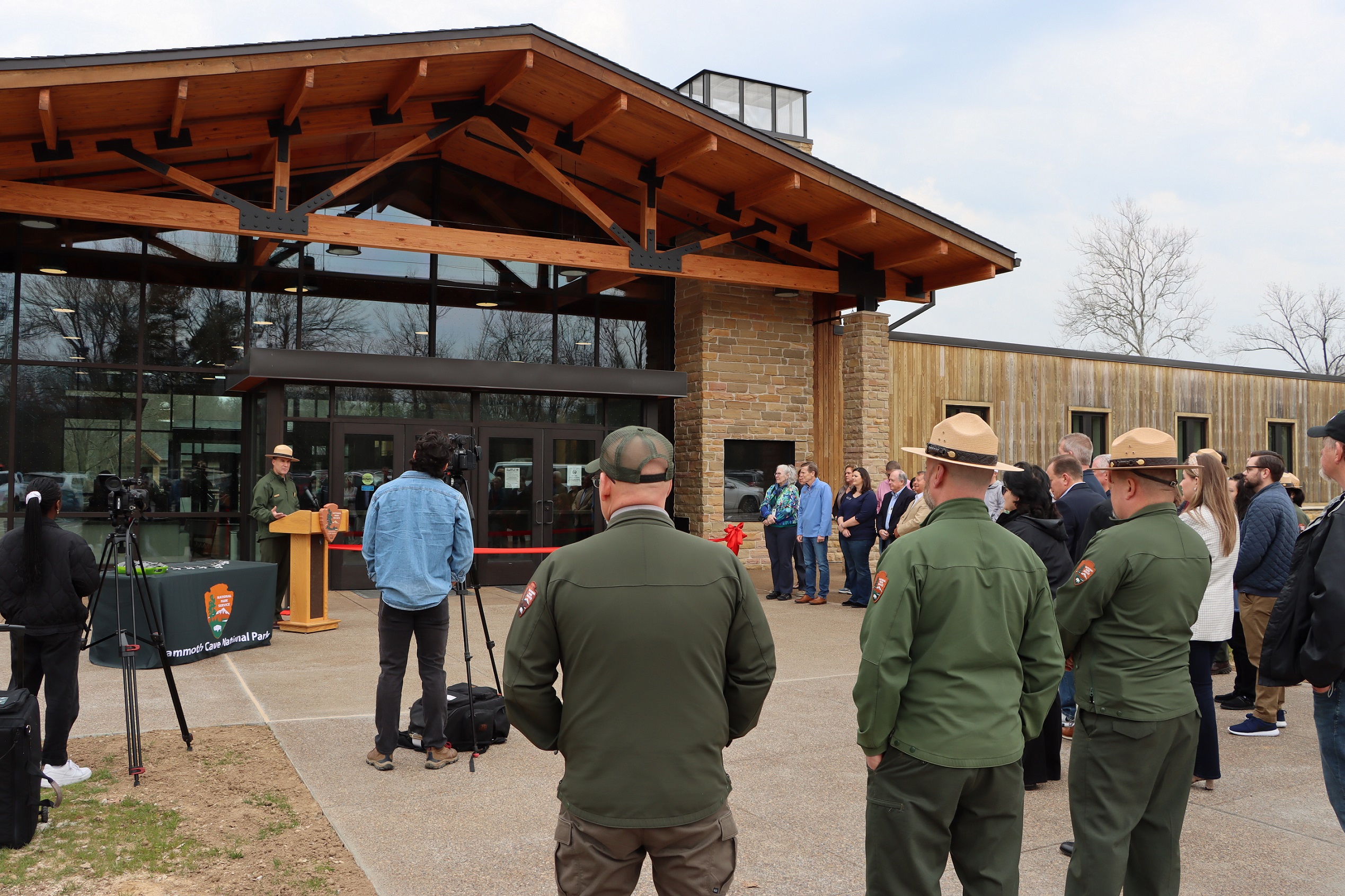 A man addresses a crowd of people standing in front of a large wood and glass building.