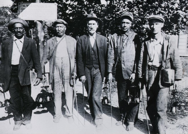 A black and white photo of five men standing next to the entrance of the cave