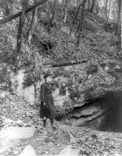 A black and white photo of a woman standing near the cave entrance.
