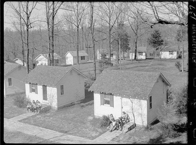 A black and white photo of several small one room cabins. People are sitting on the front steps of the buildings.