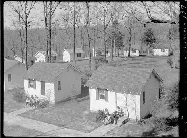 A black and white photo of several small one room cabins. People are sitting on the front steps of the buildings.
