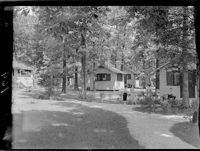A black and white photo of people gathering in front of a cabin in a wooded area.