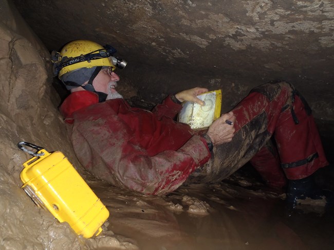 A cave researcher laying on their back in a tight cave passage while writing notes in a book.