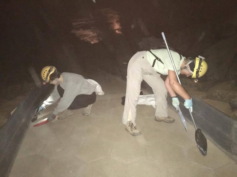 Two NSS volunteers are using brooms and dust pans to sweep up dirt, debris, and lint along the cave tour route.