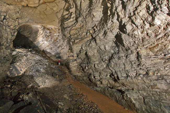 Layers of rock are seen in the wall of a cave passage. A dirt trail follows the rock wall.