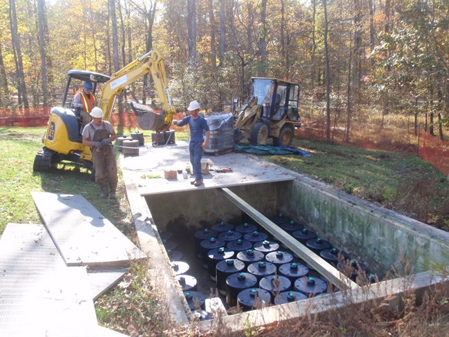 Workers use heavy machinery to change filters in a large concrete box in the ground.