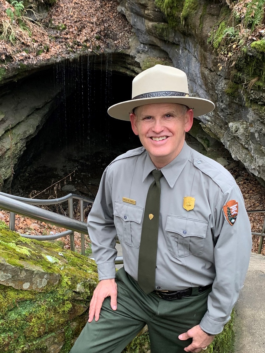 A person in a green and gray National Park Service Uniform poses for a photo in front of a large cave entrance.