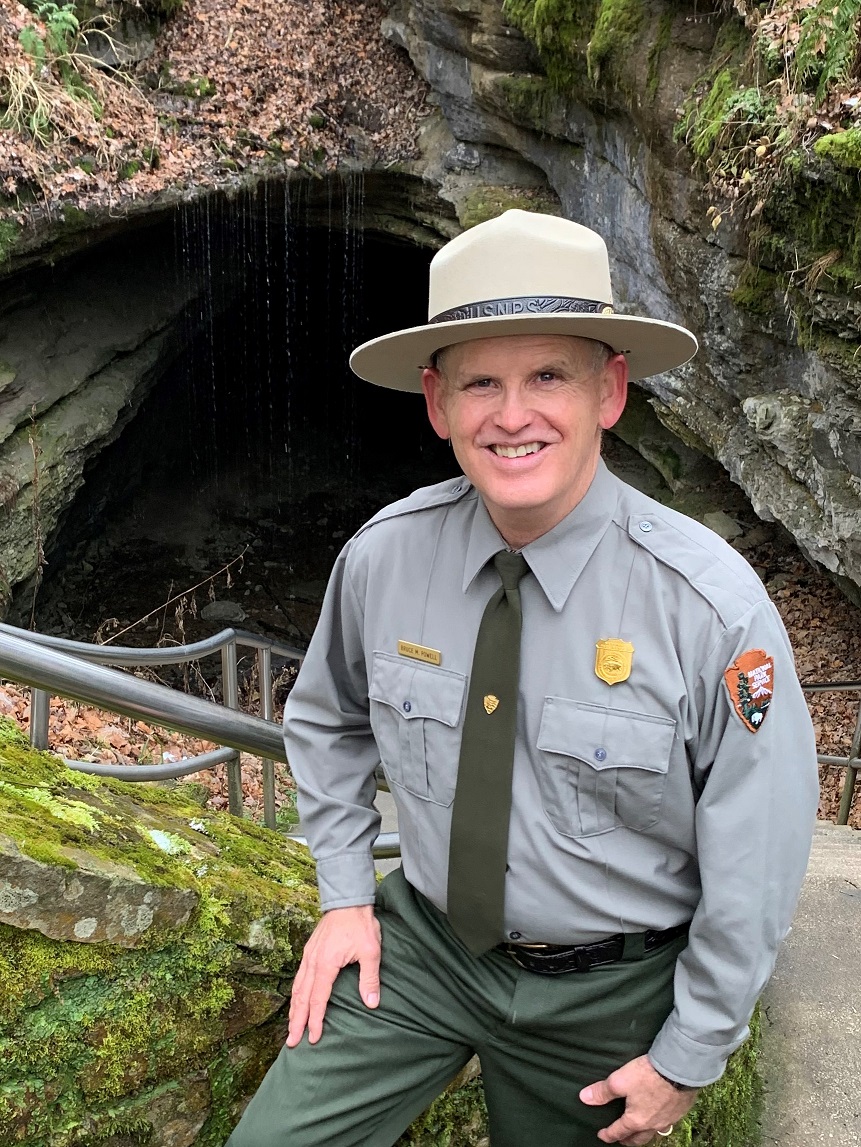 Deputy Superintendent Bruce Powell stands in front of the Historic Entrance of Mammoth Cave. A person in a green and gray National Park Service Uniform poses for a photo in front of a large cave entrance.