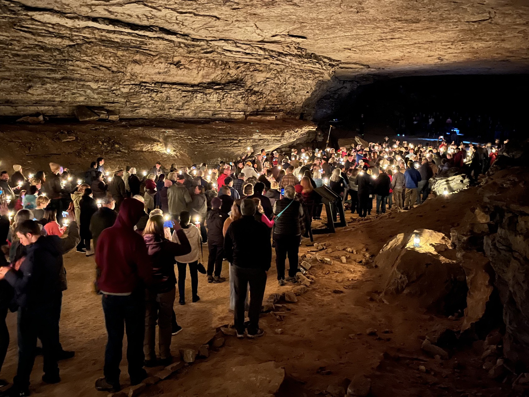 A large group of people stand in a expansive dark cave while holding small candles.