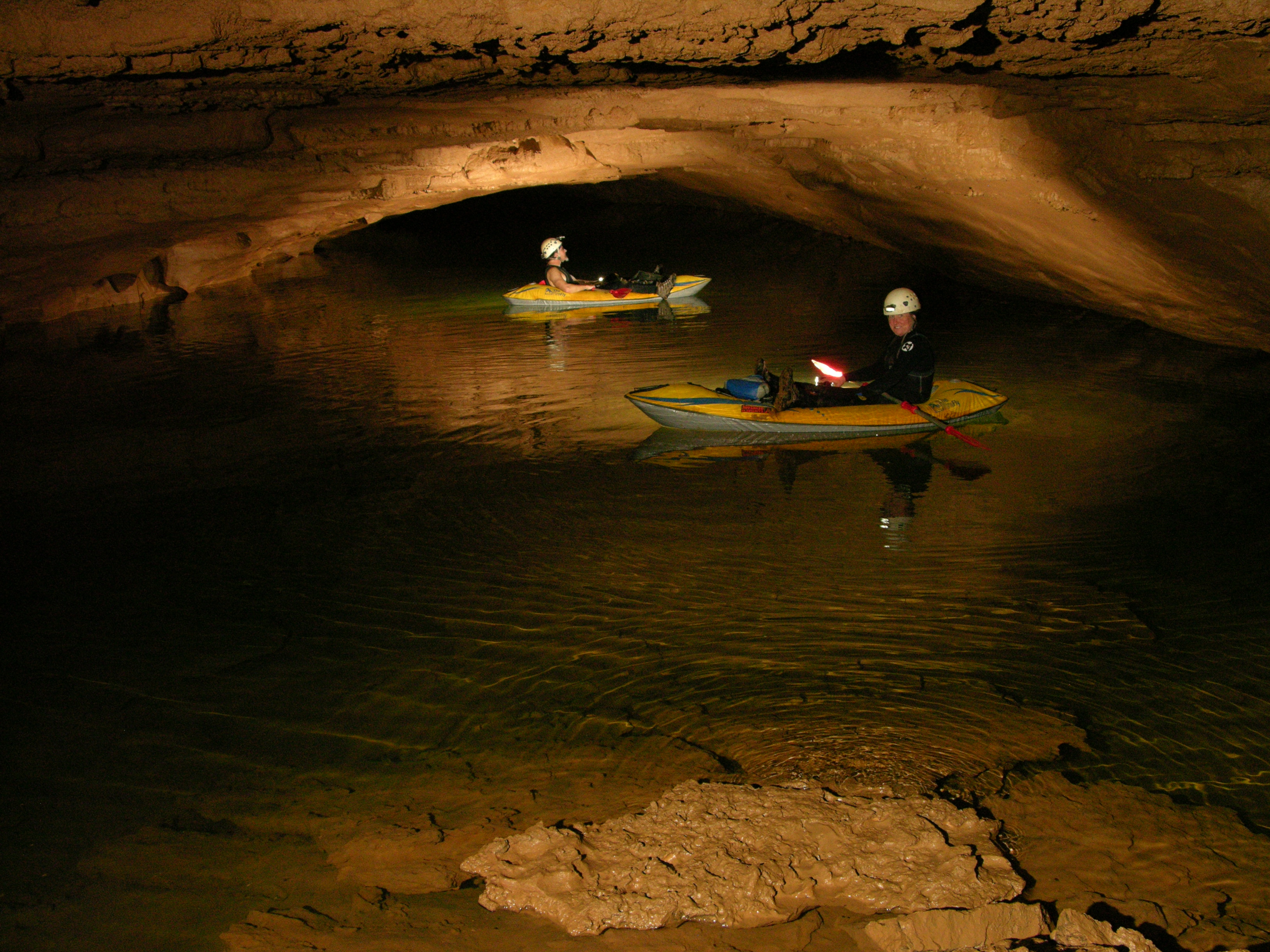 Two people wearing helmets sit in kayaks in a small body of water in a rocky cave passage.