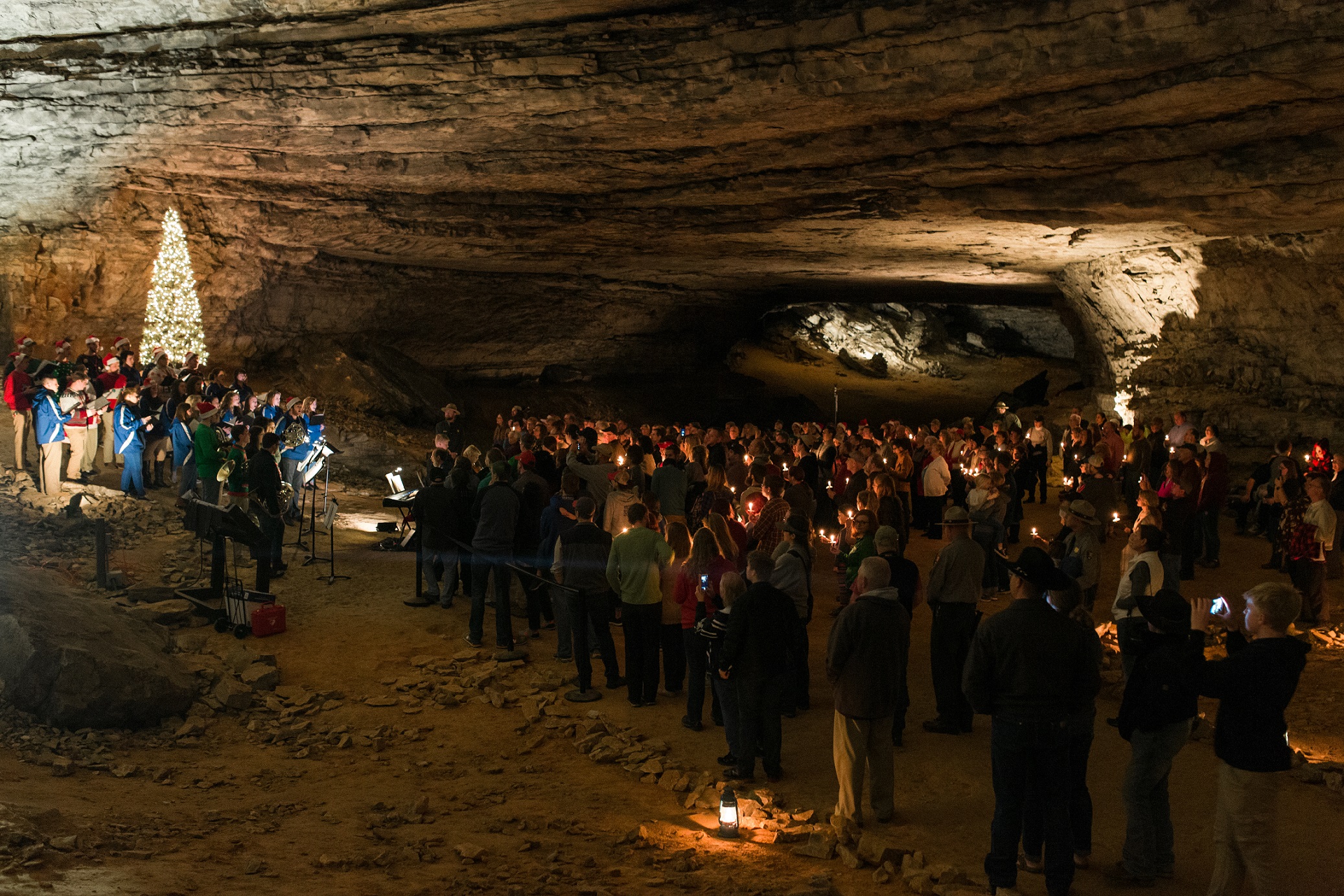 A large group of people stand as a group to watch singers on a hillside in a dimly lit cave.