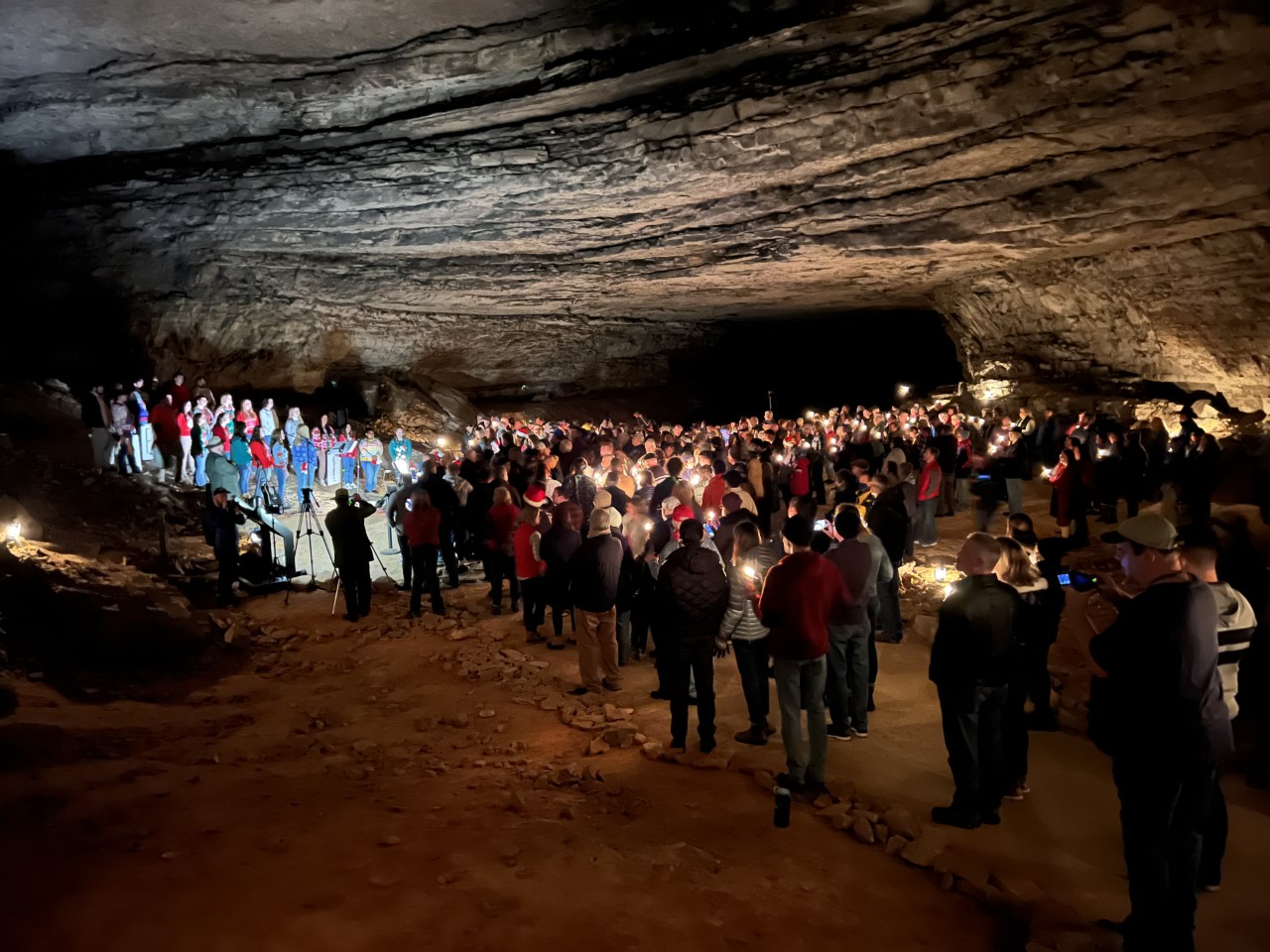 A festive crowd gathers in Rafinesque Hall inside Mammoth Cave during the annual Cave Sing event. A group of singers stand on a small hillside in front of a large crowd inside a large and dark cave.