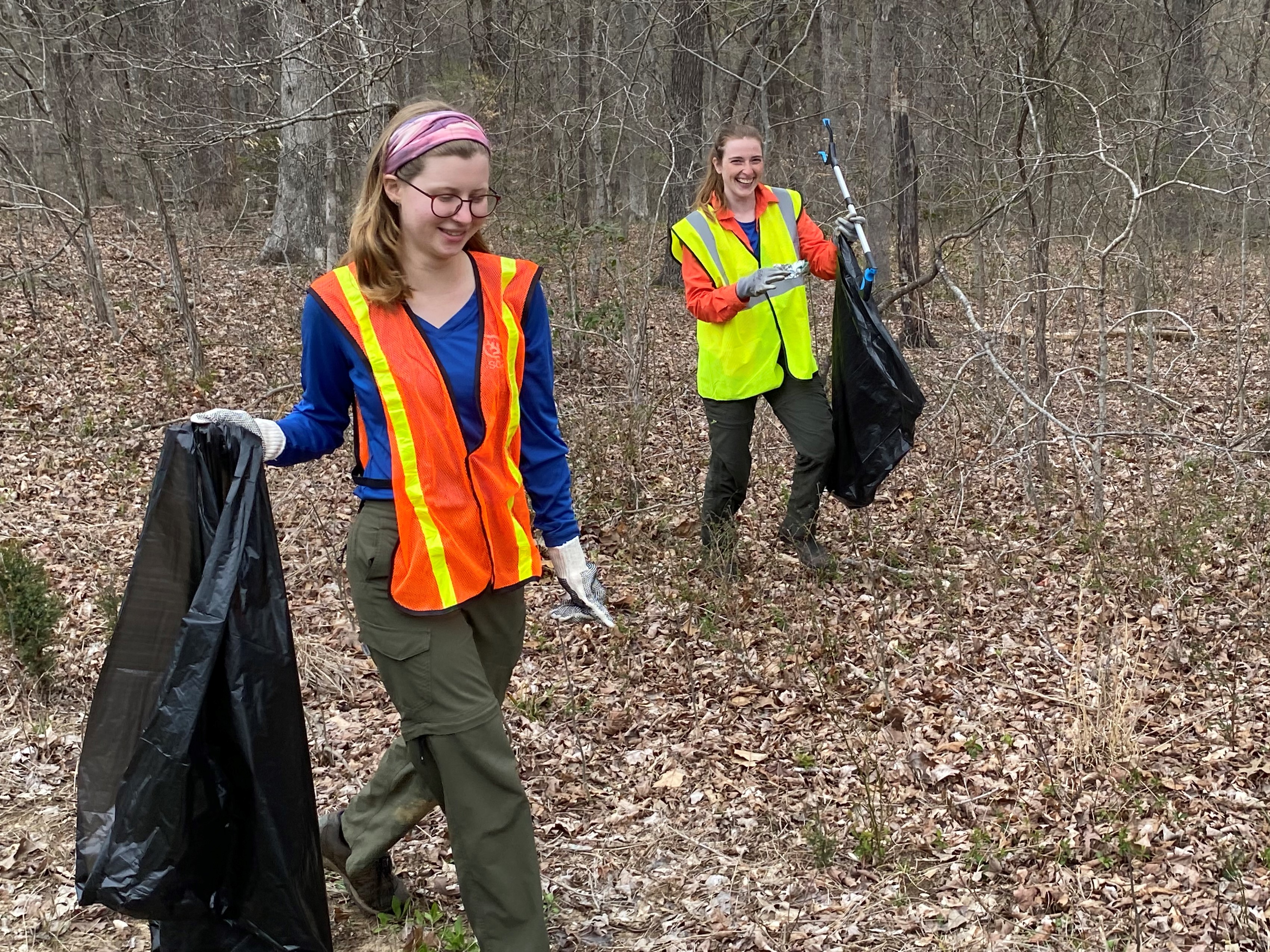 Earth Day 2023 Litter Pick Up at Mammoth Cave National Park. Two people wearing orange and yellow safety vests carry trash bags and while walking in a wooded area.