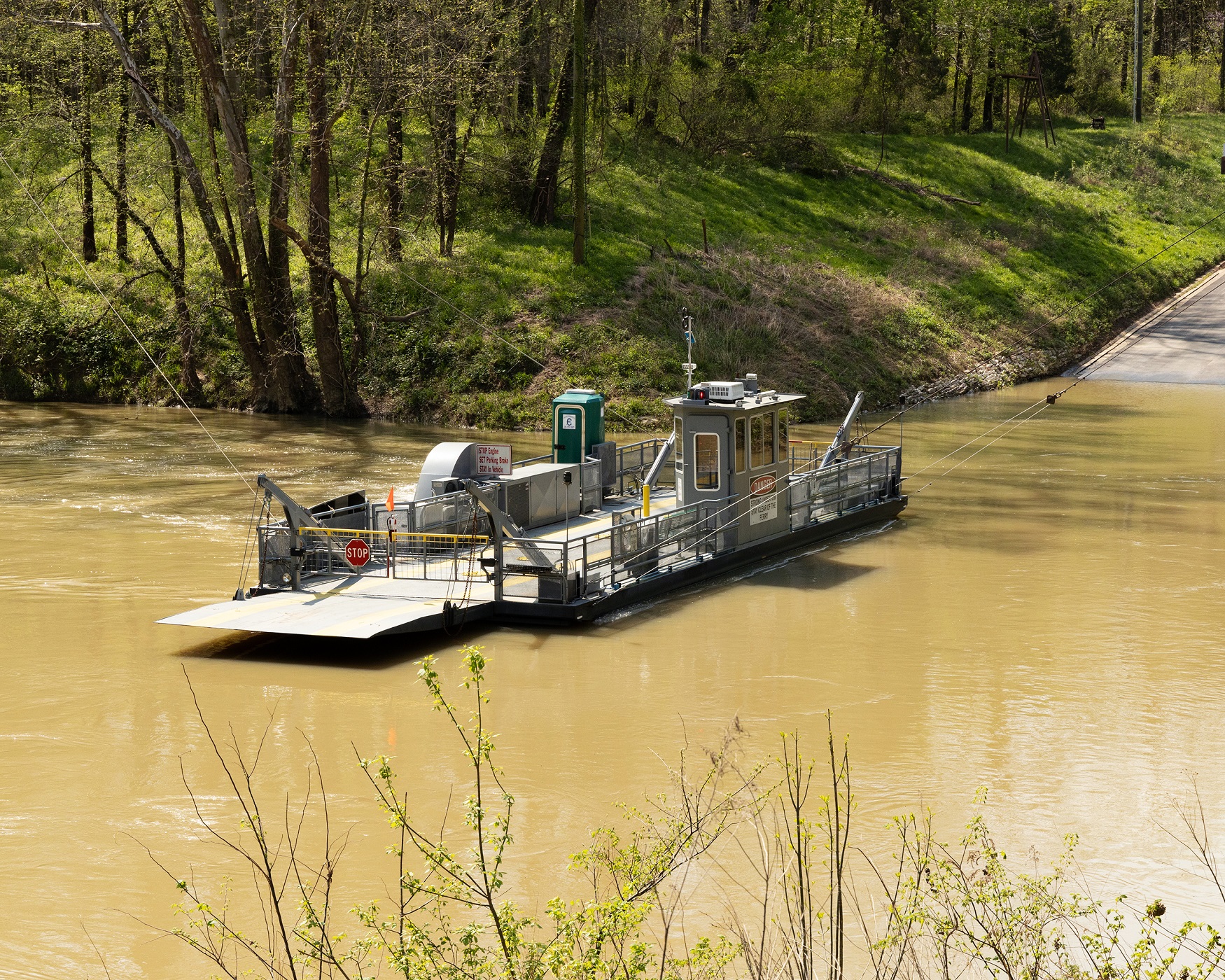 A long gray vehicle ferry boat floats in the middle of a large brown riverway