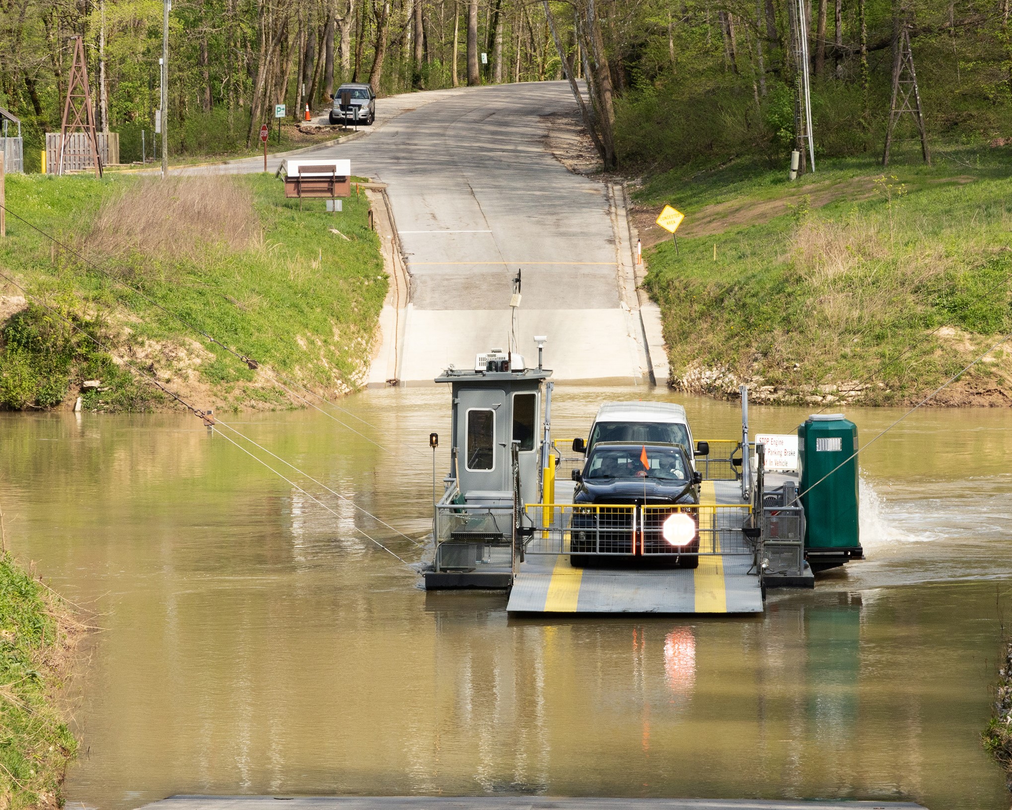 Two vehicles sit parked behind the closed gate of a small gray vehicle ferry as it moves across a muddy brown body of water.