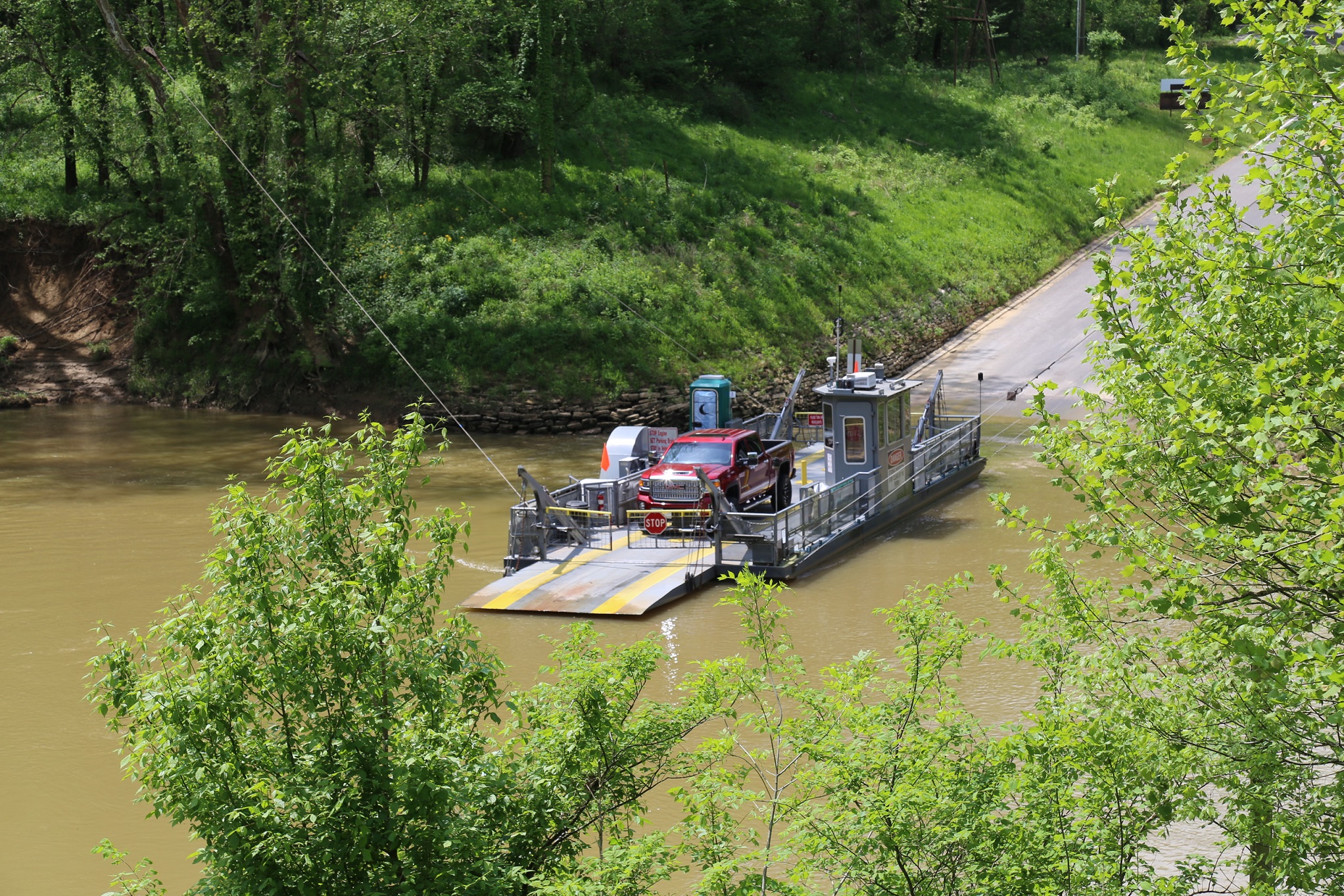 A long gray flat boat carrying a red pickup truck across a brown riverway.