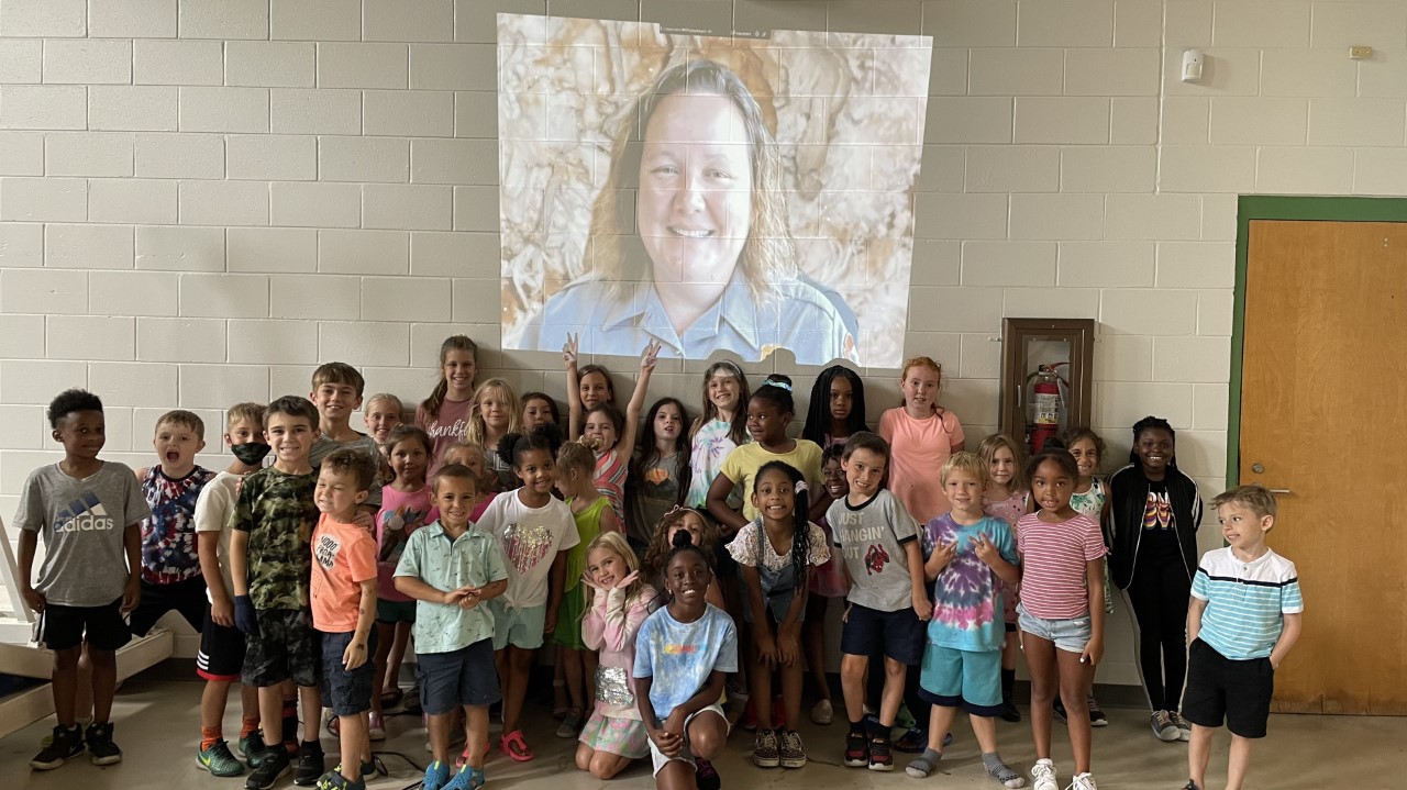 Image 2.Shackelford presenting virtually to SC group.NPS Photo A large group of children stand smiling in front of a screen displaying a person in a park service uniform.