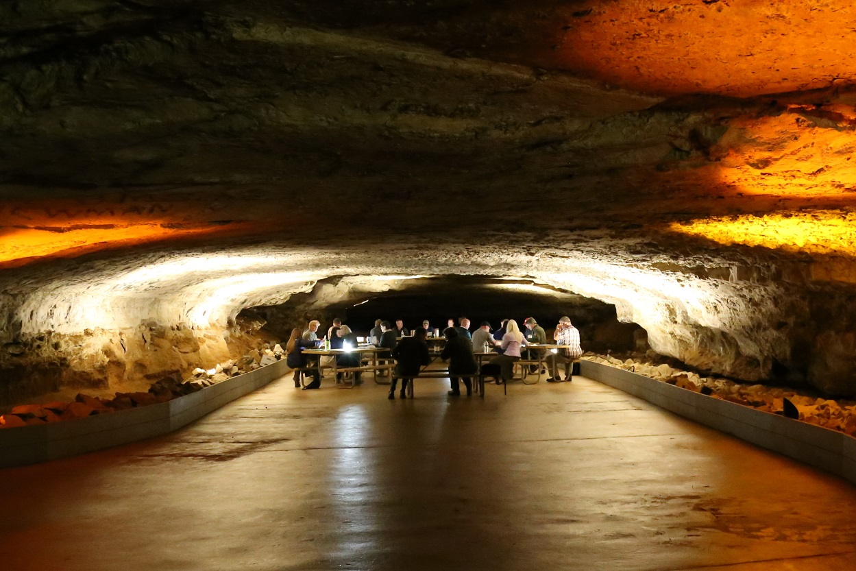 Members of the Mammoth Cave International Biosphere Reserve sit at picnic tables in the middle of a wide oval shaped cave passage illuminated by white and orange lighting.