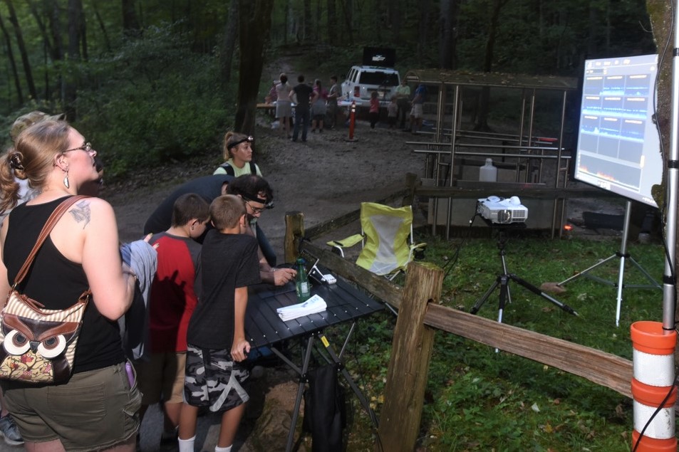Visitors are able to see different bat monitoring techniques during Bat Night including equipment that monitors echolocation. A family stands in front of a display screen in a wooded area.