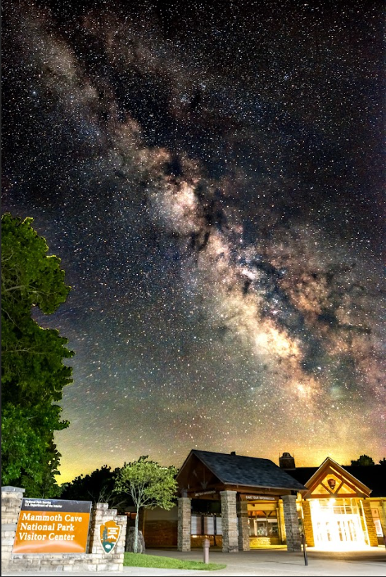 MACA Visitor Center with Milky Way.Jacob Corder Photo A cloudy haze of stars in a dark night sky is visible above a large building and a sign that reads “Mammoth Cave National Park Visitor Center”.