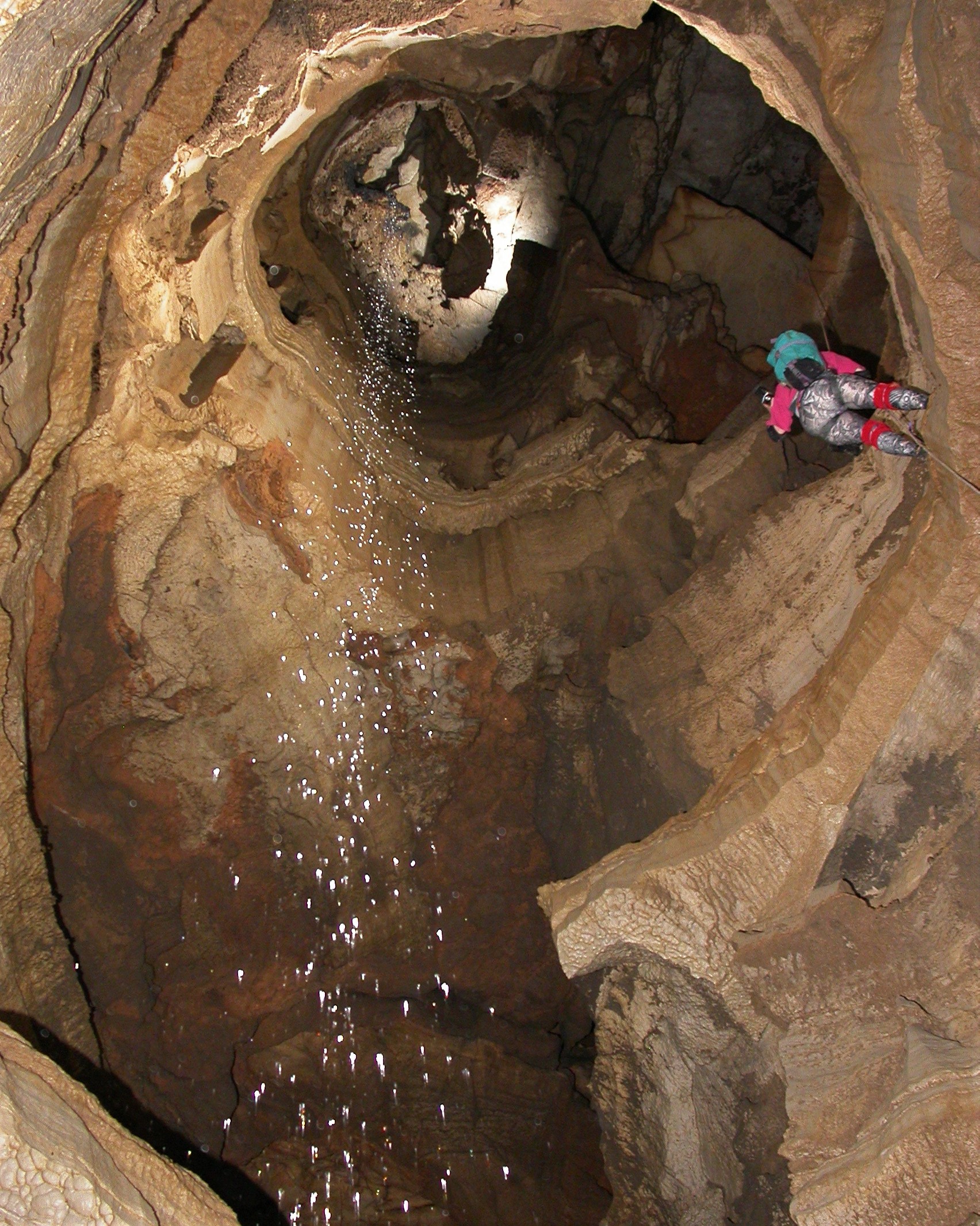 A person holds herself up on a long rope in a rocky, jagged cave with water streaming next to her.