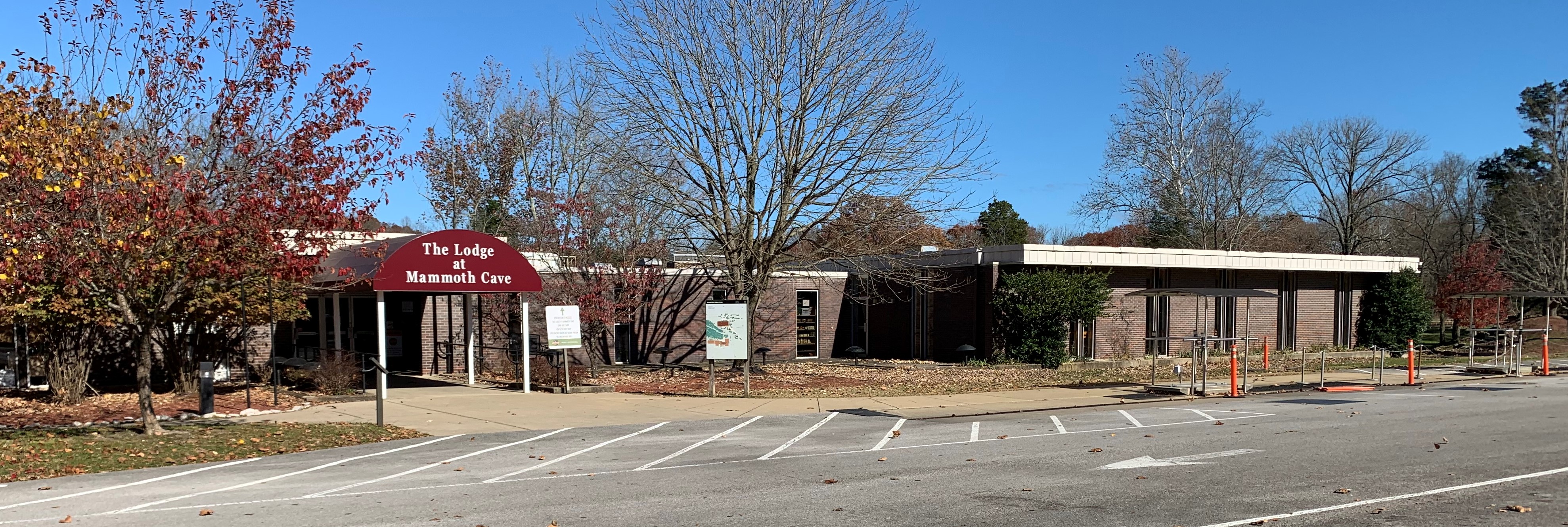 A long one story brick building with a dark red awning that reads "The Lodge at Mammoth Cave" stands behind leafless trees with bright blue sky above.