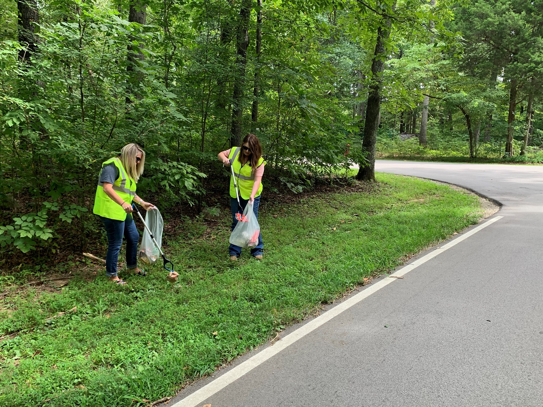 Two people in bright yellow vests use long poles to pick up paper and plastic trash items in a wooded area next to a road.