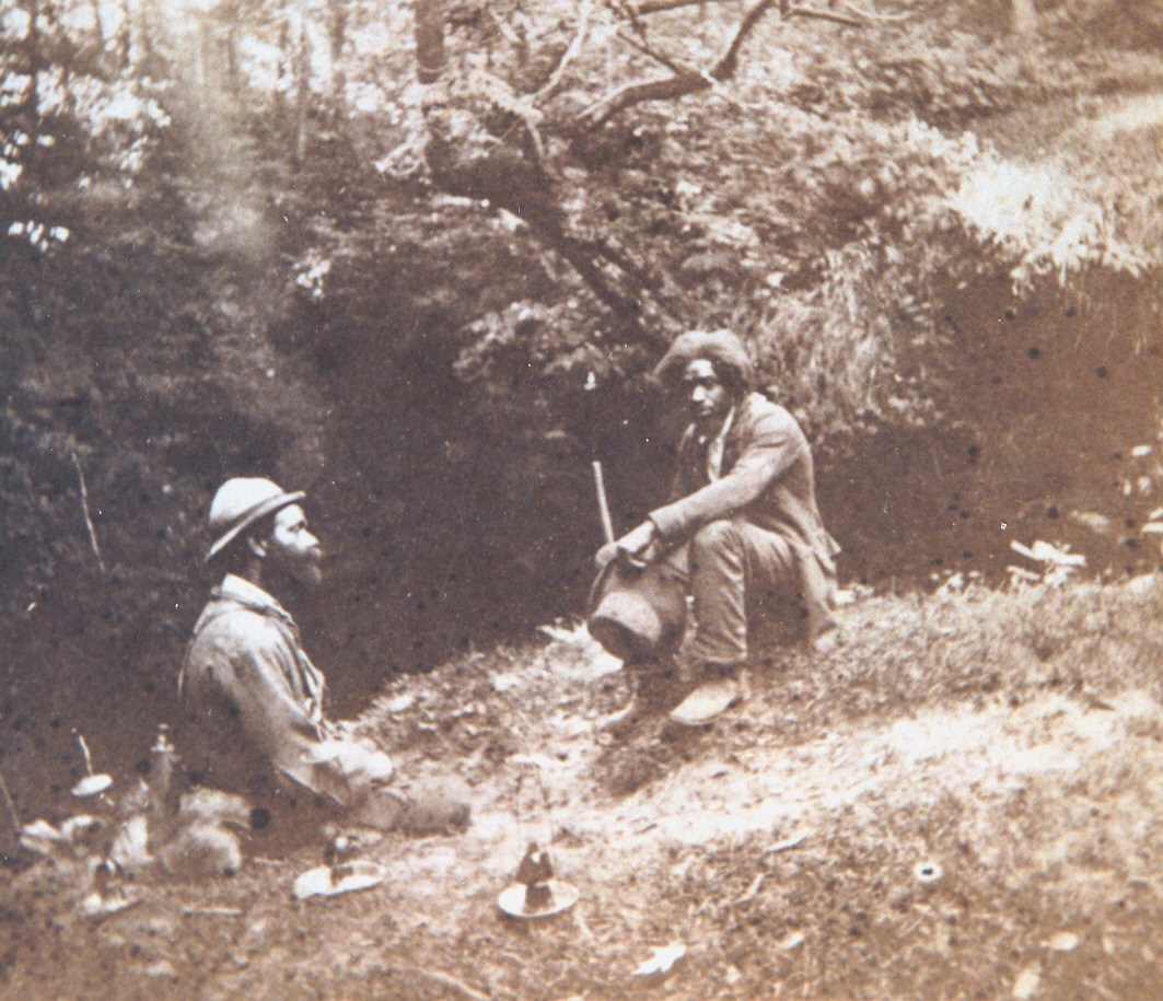 Black and white photo of two men sitting on a grassy hillside next to a cave entrance.