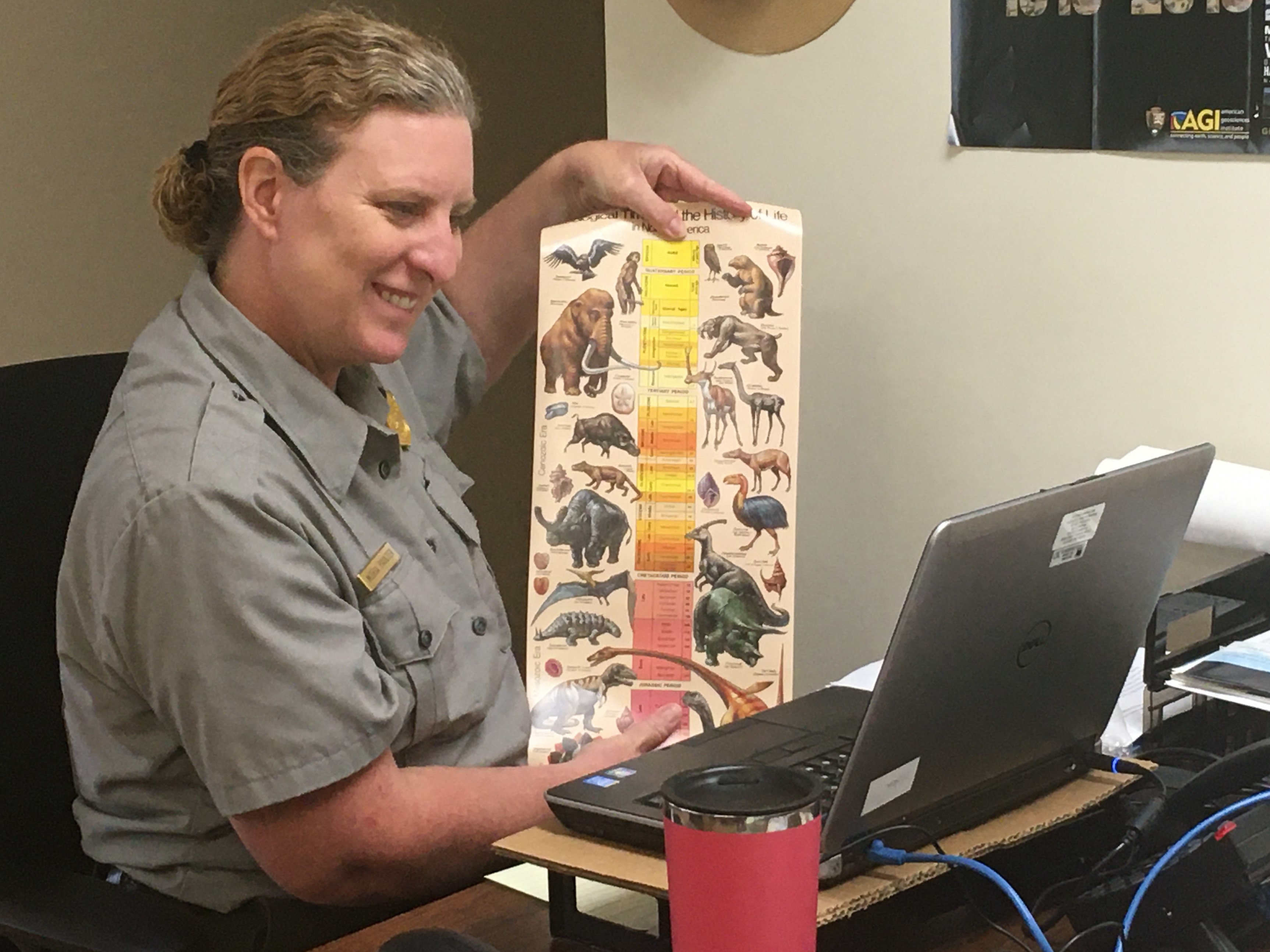 A virtual environmental education program is conducted at Mammoth Cave National Park. A woman in a park ranger uniform holds up a chart for her computer screen that show prehistoric mammals and dinosaurs