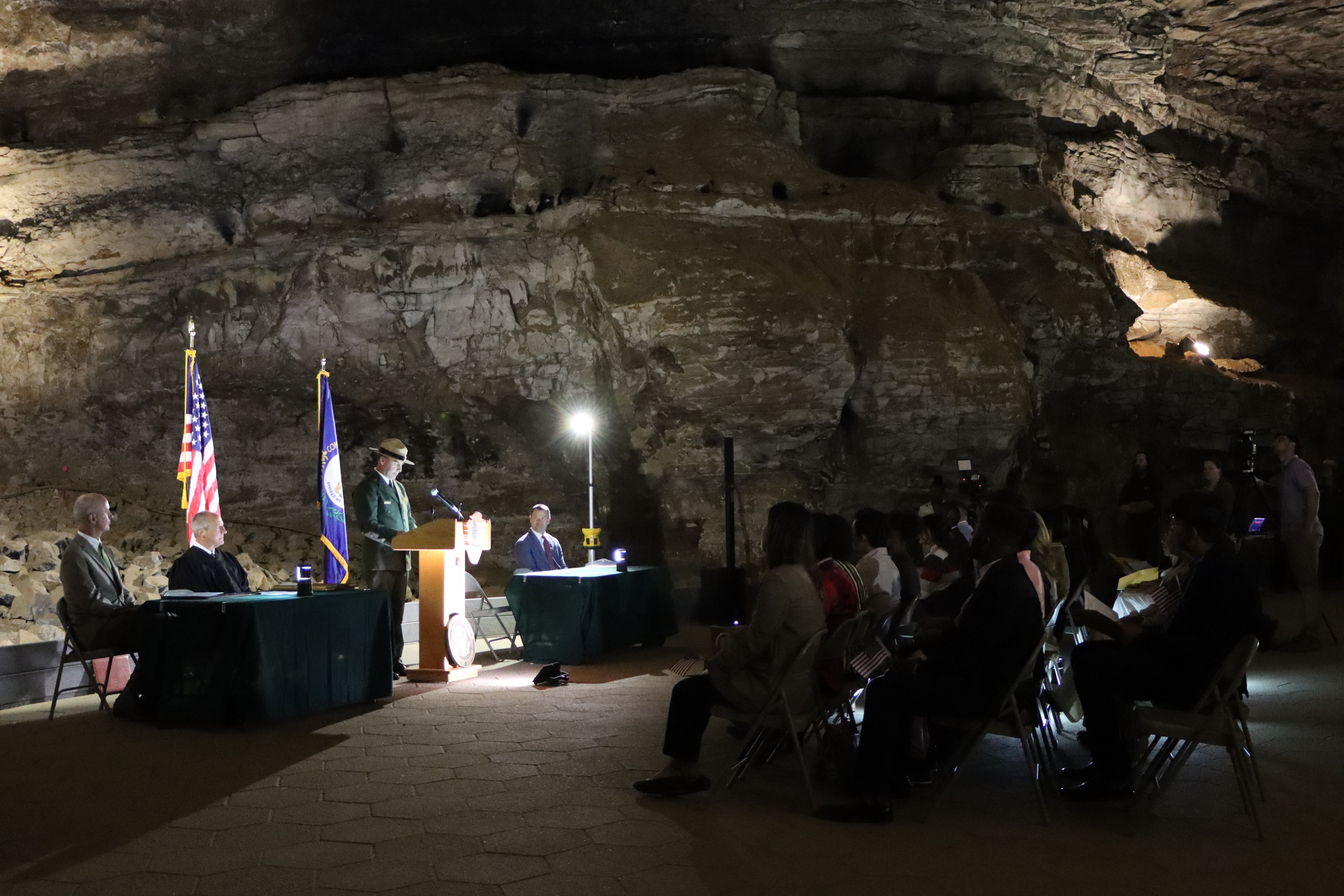 A man in a National Park Service Uniform stands at a podium in front of a seated crowd of people inside a dimly lit cave.