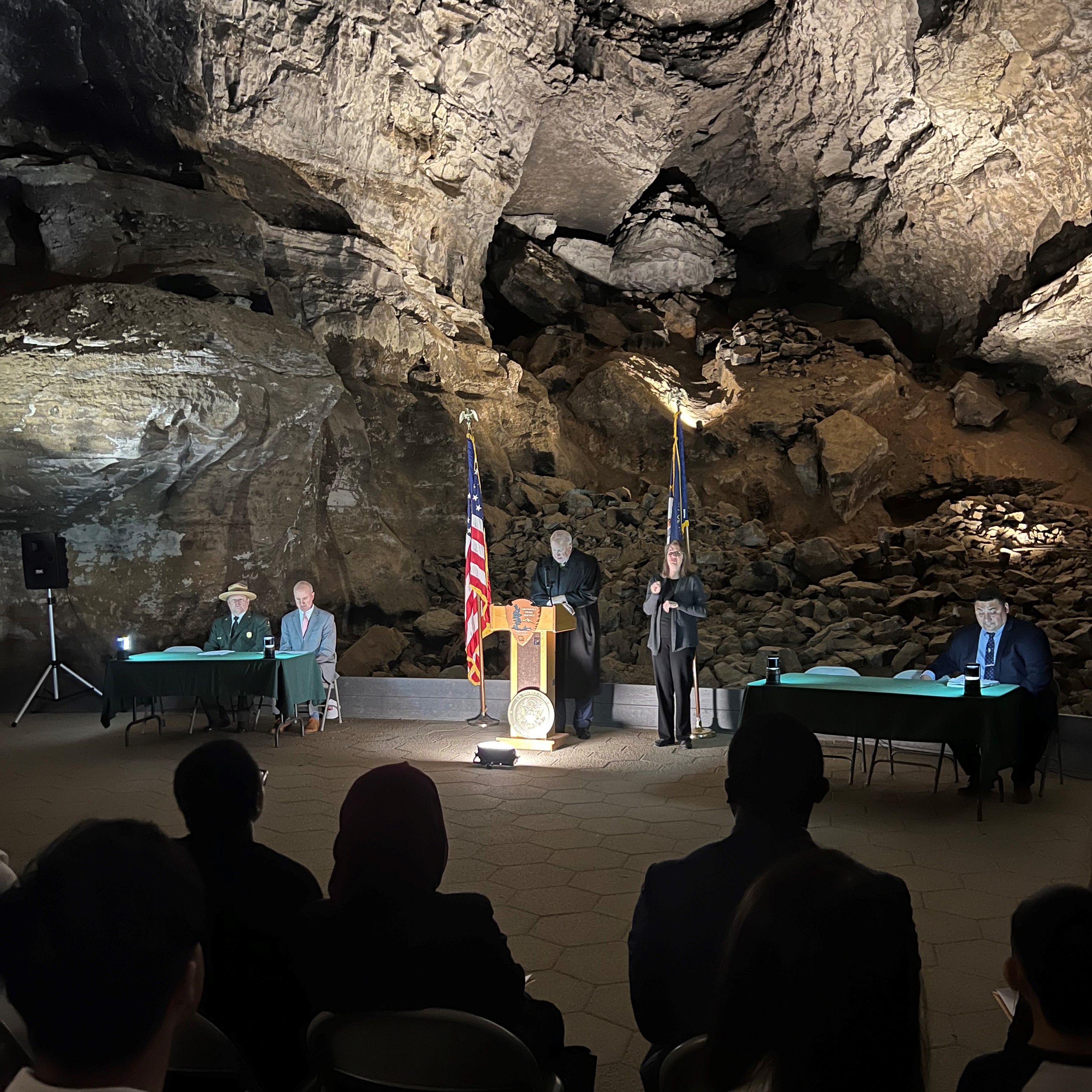 A judge wearing a black robe stands behind a podium and next to the American and Kentucky state flag in a rocky cave.