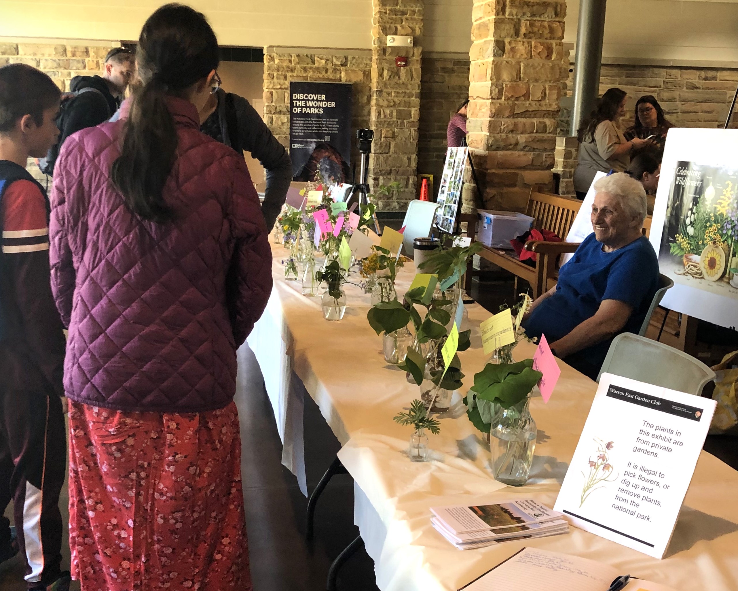 A woman sits behind a long table with clear glass vases containing various shaped green plants displayed on top.